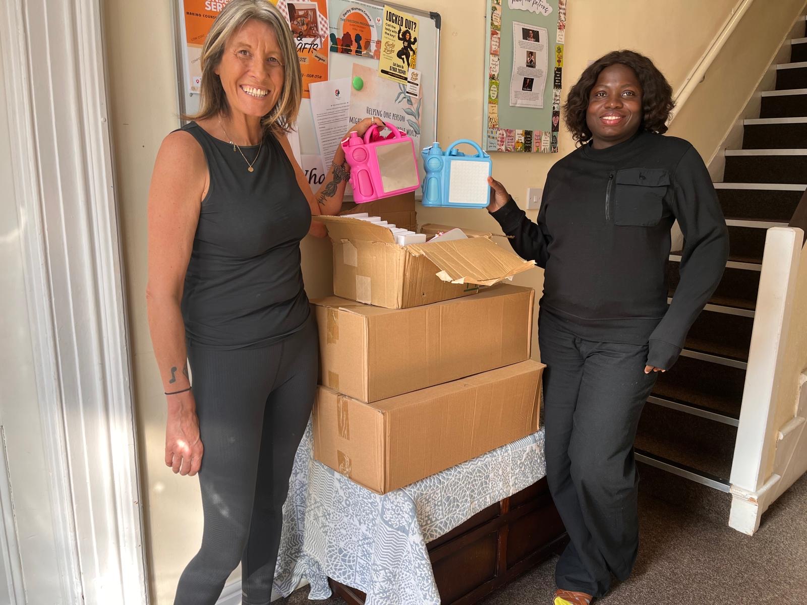 Two women at Derby Women's Centre holding pink and blue lunch boxes beside boxes of donated school packs